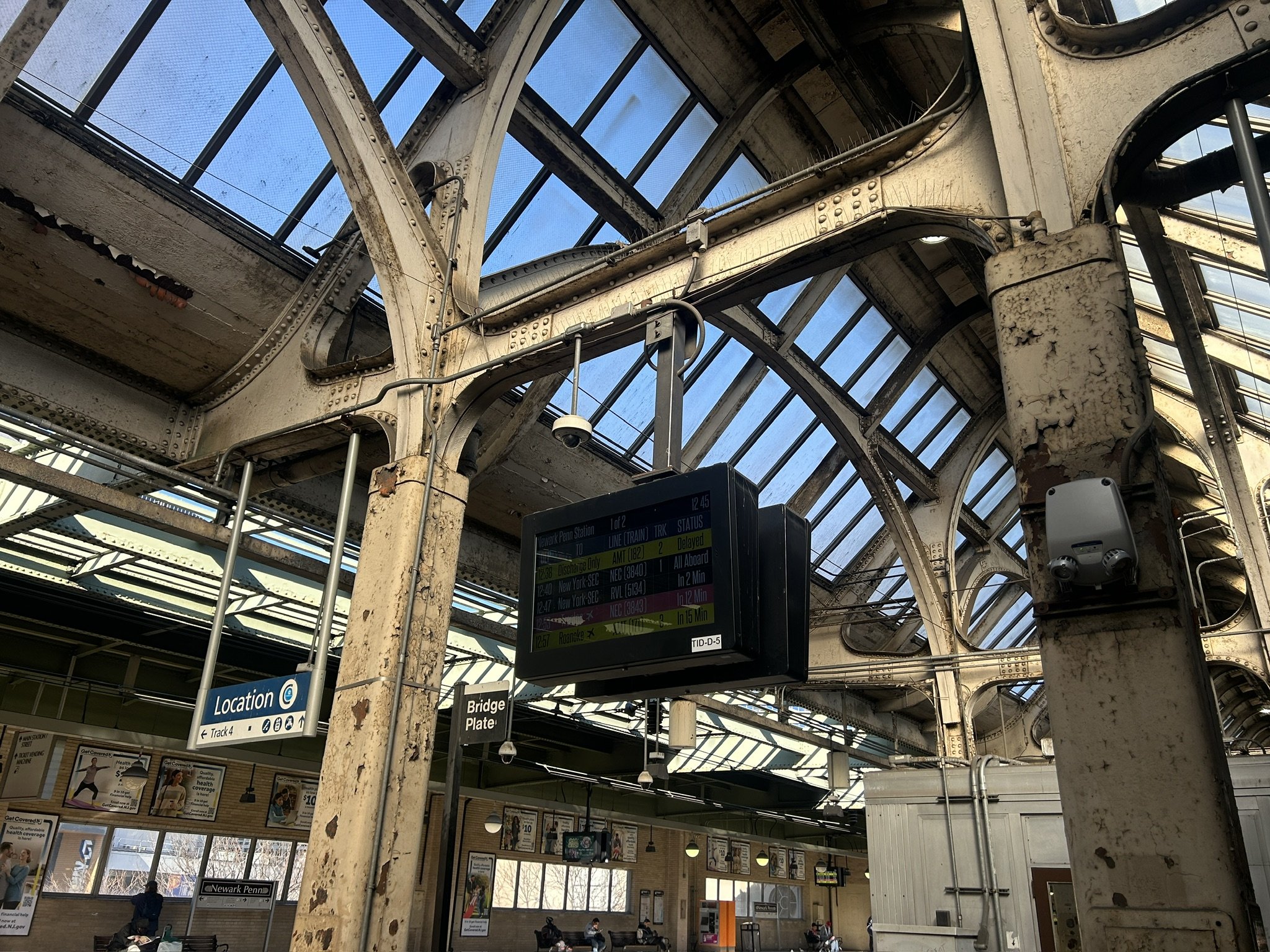 A textured image of rusting steel beams and steel framed skylights at Newark Penn Station with a visual history of different improvements and technology added over time including an electronic time schedule on a video screen.