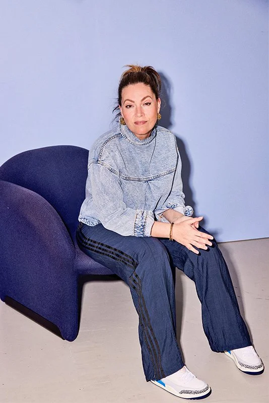 Ilse sitting on a dark blue chair, wearing a light blue denim jacket, dark blue track pants with black stripes, white sneakers, gold earrings, and a bracelet, against a light blue wall.