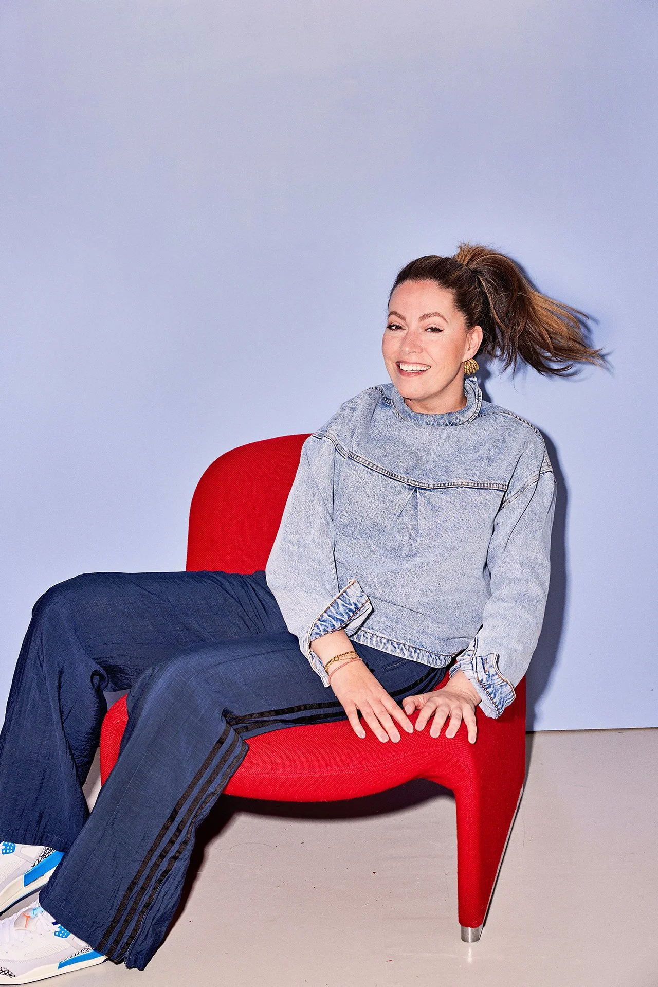 A woman with a ponytail hairstyle, wearing a denim jacket and dark pants, sitting on a red chair and smiling. The background is a plain light purple wall.