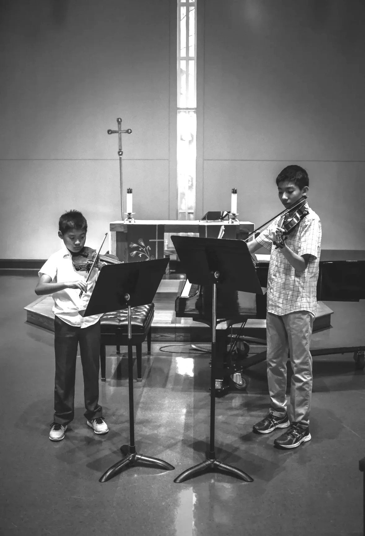 Two boys playing violins in a church or concert hall, standing in front of music stands with candles and a cross on the altar behind them.