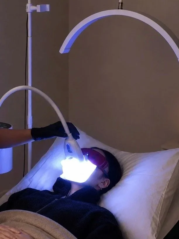 A person lying on a bed receiving a bright LED facial treatment from a healthcare professional wearing black gloves. The person is wearing protective goggles, and the device emits a strong light onto their face.