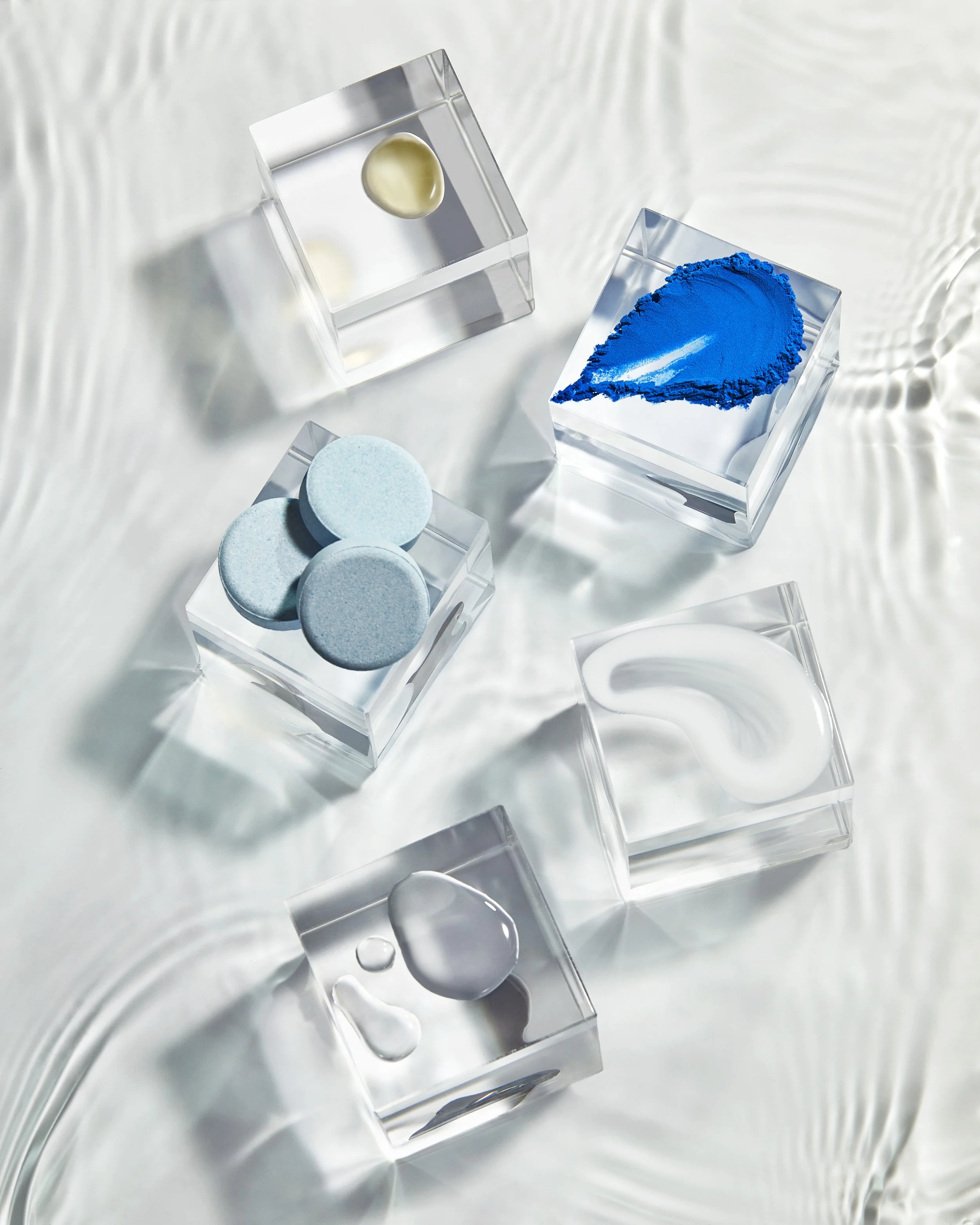 A clear glass container holding blue powder, a clear container with light blue tablets, and a clear container with a round yellow pill, all placed on a metallic surface.