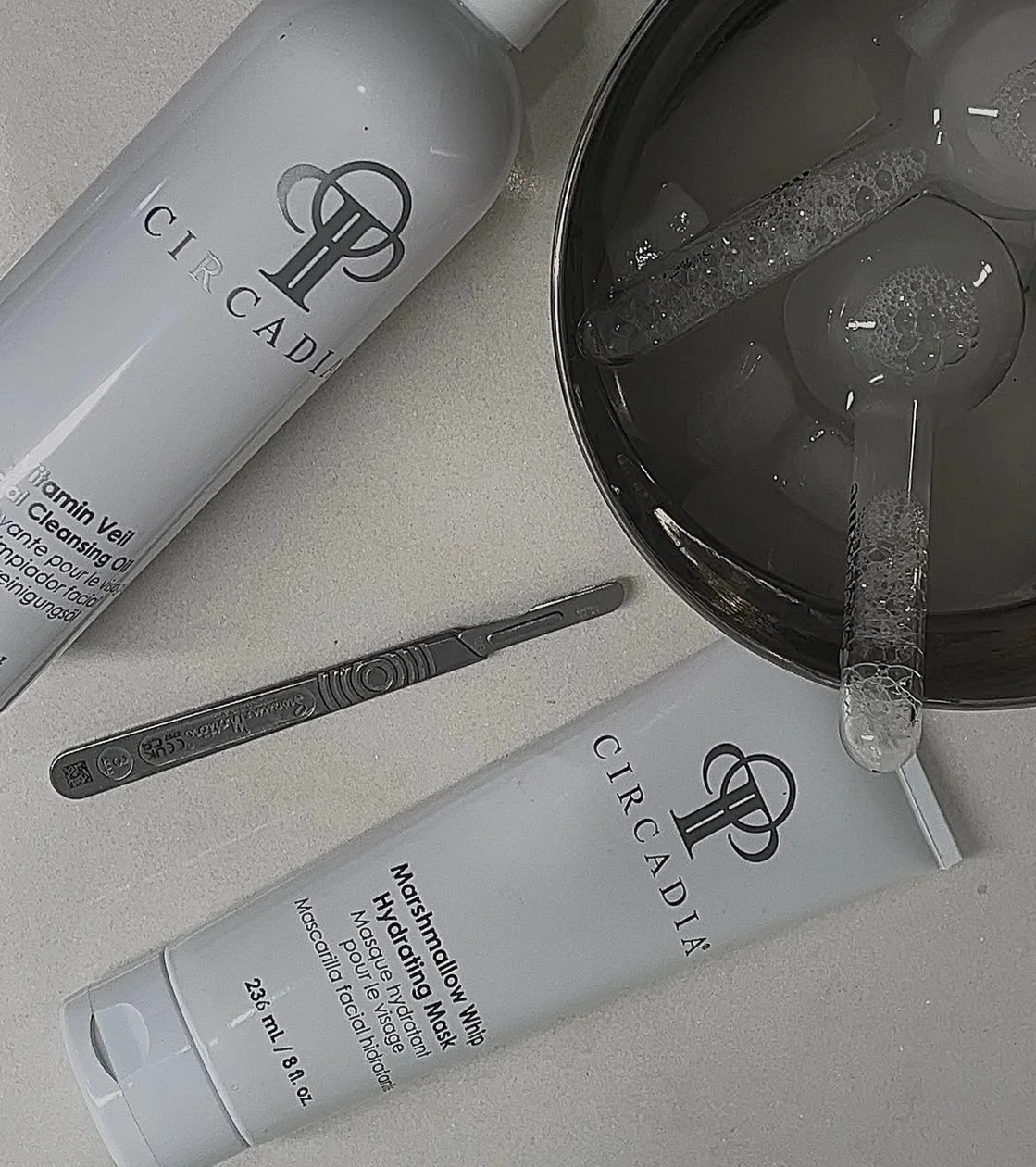 Cirqadía facial cleansing products, a tube of hydrating mask, a glass bowl with facial tools, and a black spatula on a white surface.