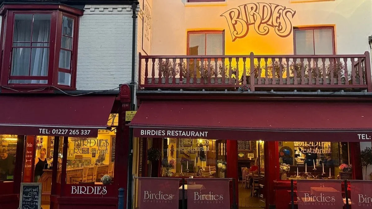 Exterior view of Birdies Restaurant with a maroon and yellow facade, signage displaying the restaurant's name, windows showing the interior, and a balcony on the upper floor with potted plants.