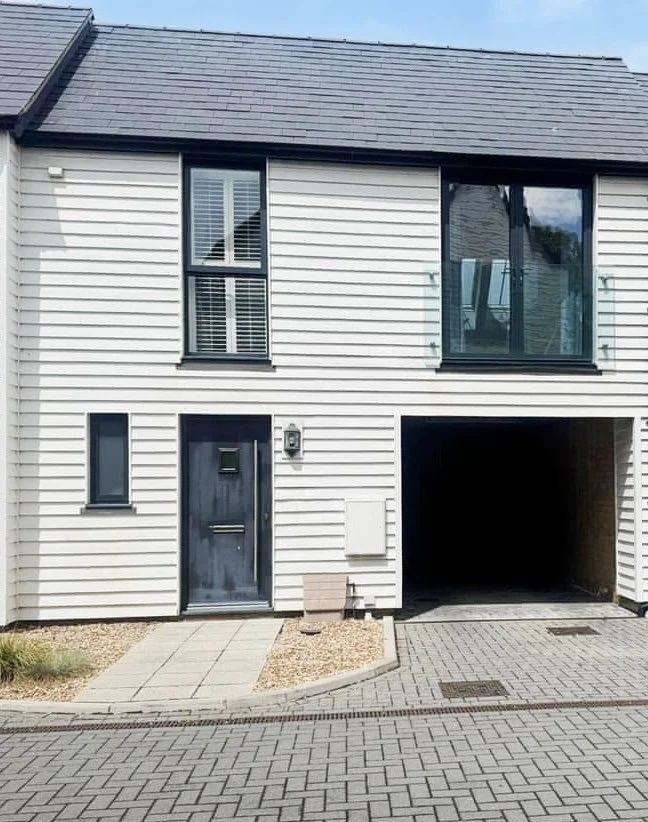 Front of a modern two-story house with beige siding, a black front door, a small window, and a large garage opening. There is a small pathway and a gravel area beside the door.