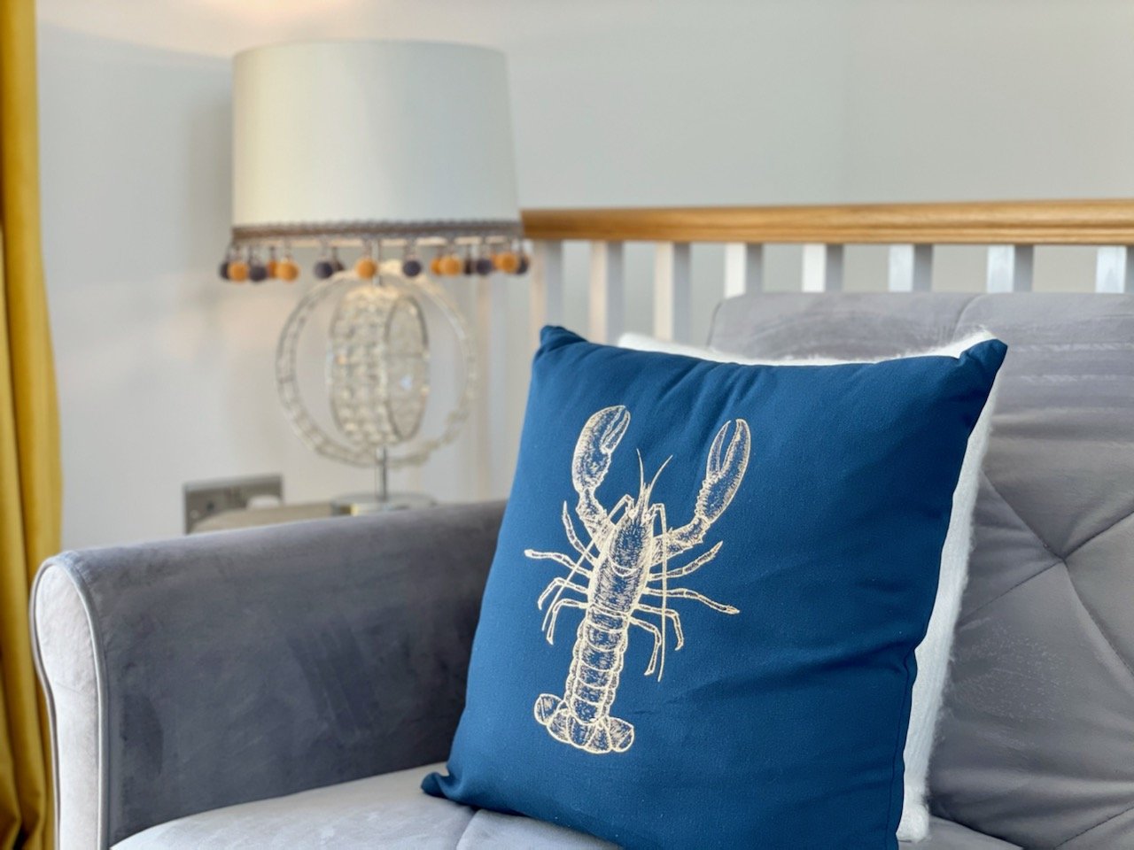 Blue throw pillow with a white lobster illustration on a bed, guest room decor with a gray headboard and a white lamp with a beaded shade in the background.
