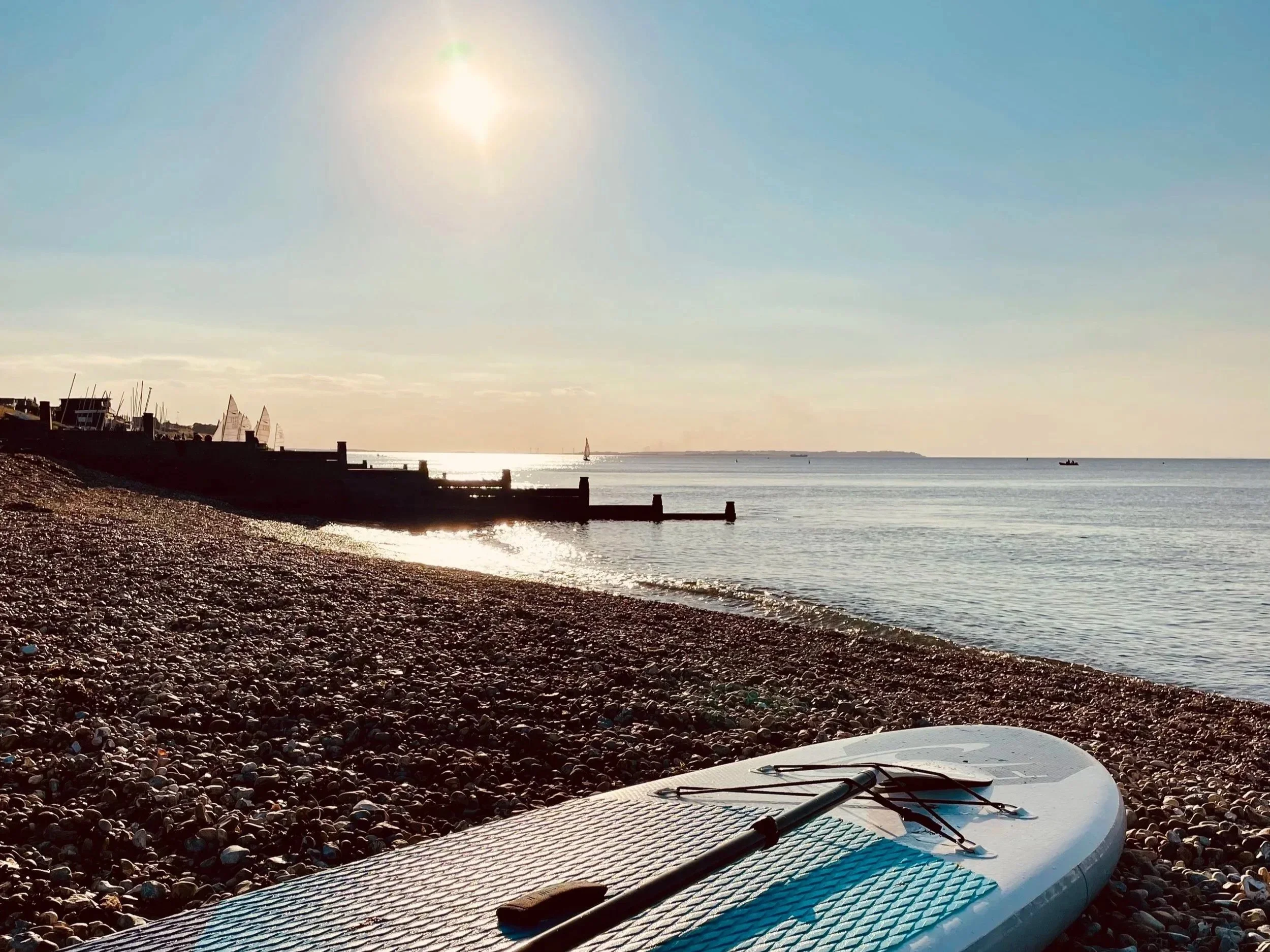 View of a pebble beach with a paddleboard lying on the shore, sailboats in the water, and a boat in the distance under a clear sky with the sun shining brightly.