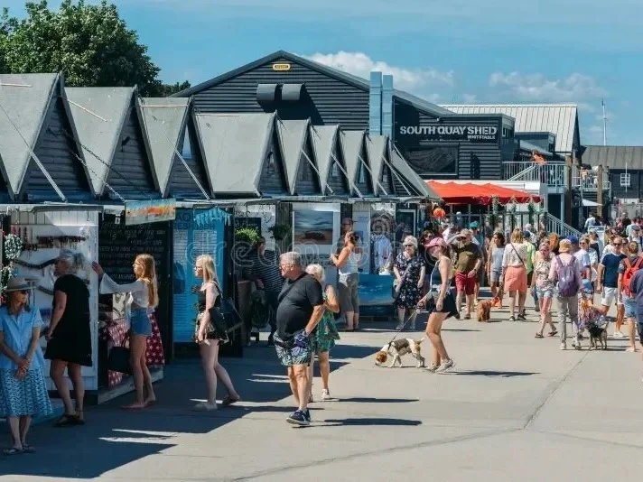 People walking and shopping at an outdoor market with shops and food stalls, including a building labeled 'South Quay Shed' in the background, under a blue sky with some clouds.