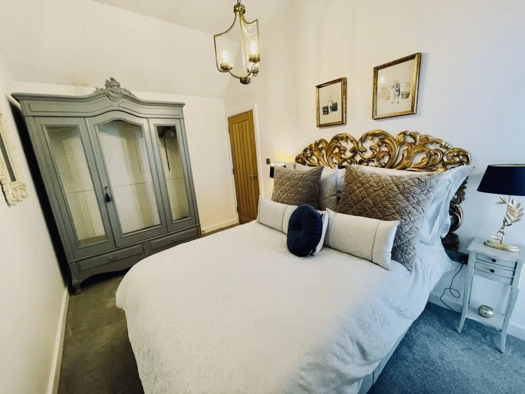 A bedroom with a large bed featuring an ornate gold headboard, topped with multiple decorative pillows, a gray wardrobe in the corner, a small white nightstand with a black lamp, and framed artworks on the wall.