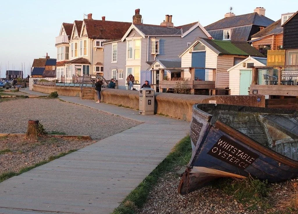 Beachside walkway with colorful houses and boats, including a boat labeled 'Whitestark Oysters' in the foreground.