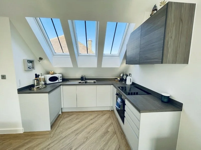 Modern kitchen with white cabinets, gray countertops, and three skylight windows showing a blue sky with some rooftops and chimney outside.