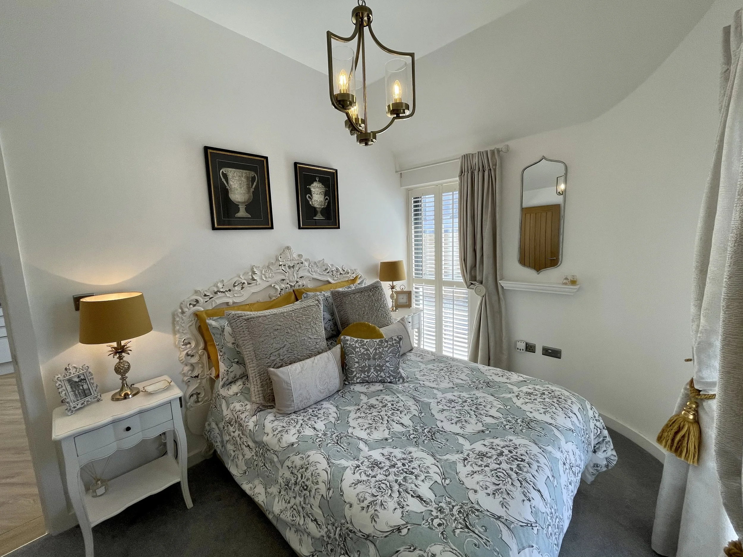 A bedroom with a decorative white bed frame, gray and white patterned bedding, and multiple pillows. There is a small white bedside table with photo frames, a lamp, and decorative items. The wall above the bed has two framed artwork pieces of classical urns. A window with white plantation shutters and beige curtains is on the right side. A brass chandelier with glass shades hangs from the ceiling, and a wall mirror and fireplace are partially visible.