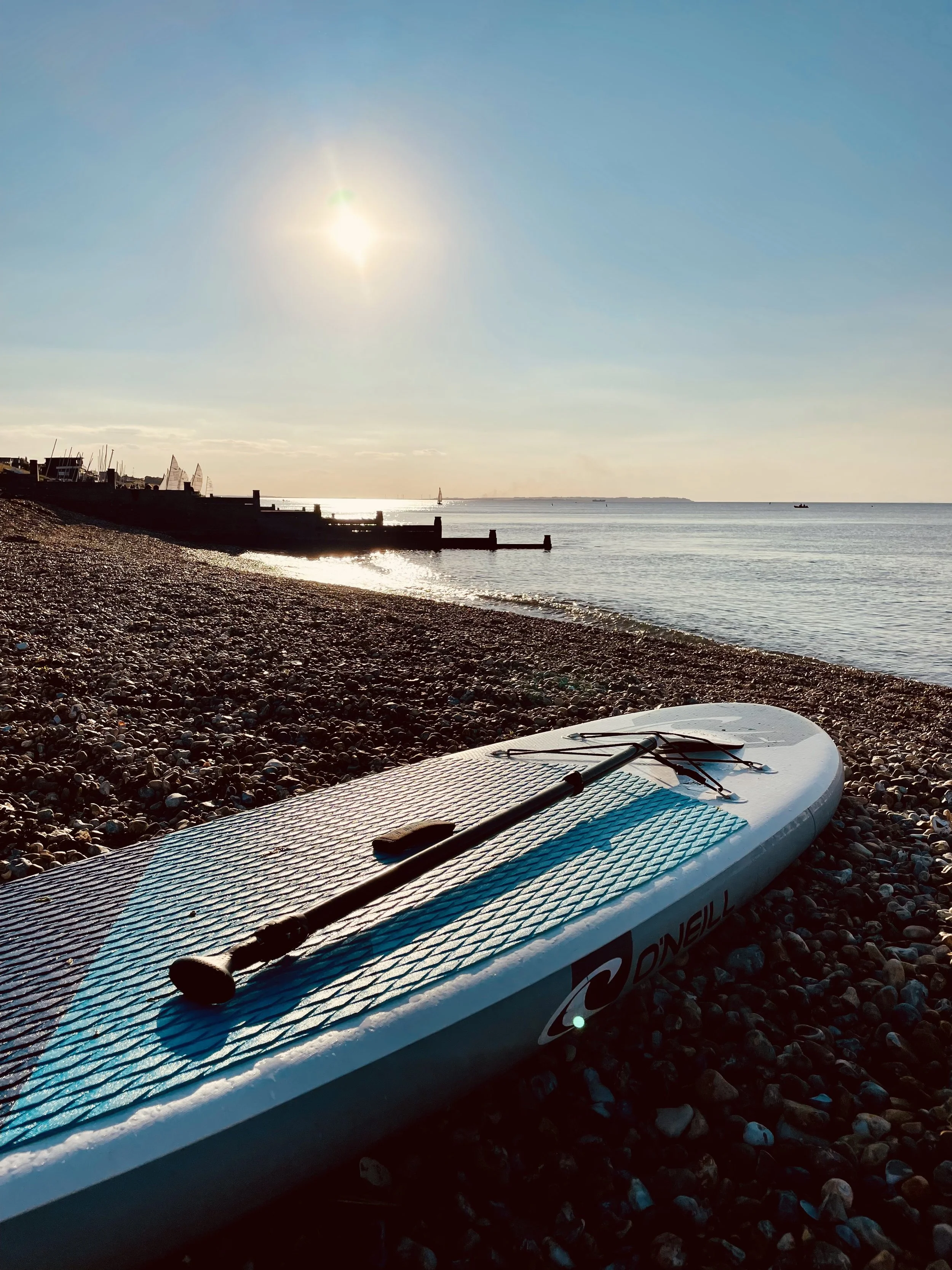 A paddleboard resting on a pebbled beach with a paddle on top, overlooking calm water under a bright sun.