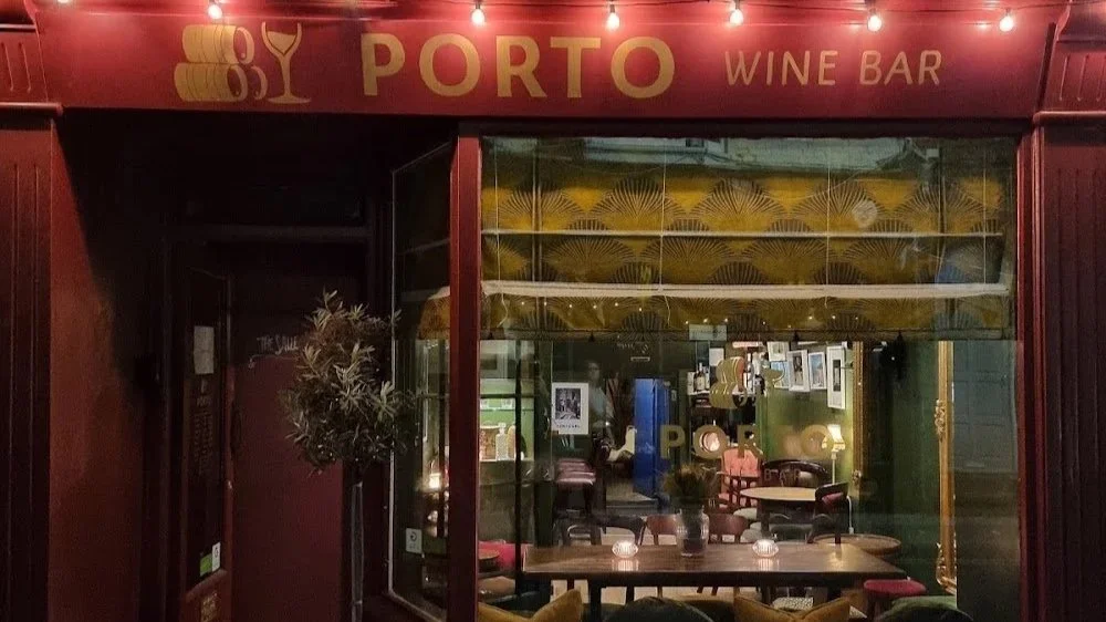 Outside view of Porto wine bar with illuminated sign and inside tables visible through large window, decorated with hanging lights and wall art.