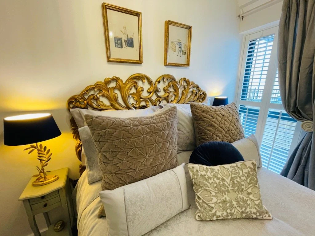 A bedroom with a white bed, decorative pillows, and an ornate gold headboard. There are two black lamps with gold bases on side tables, and framed artwork on the wall above the bed. A window with blinds and curtains is on the right side.