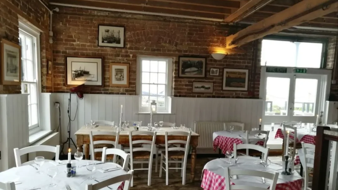 Cozy restaurant interior with brick walls decorated with framed artwork, white wooden chairs, tables with white tablecloths, some with red and white checkered tablecloths, candlelit table settings, large windows allowing natural light, exposed wooden beams on the ceiling, and a rustic, inviting atmosphere.