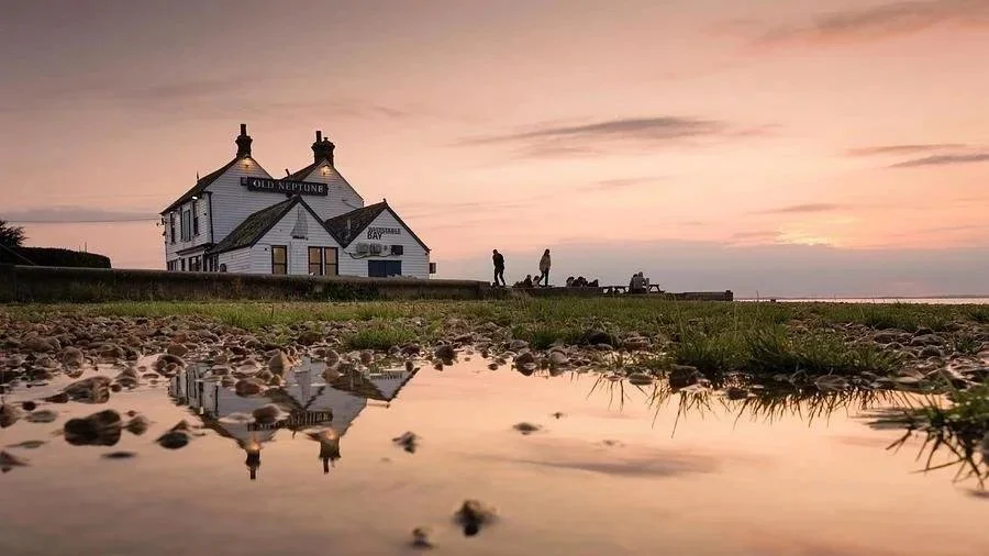 A lighthouse with the sign 'Old Neptune' on it, with a silhouette of people walking along the shore at sunset, and their reflection visible in a small pool of water.