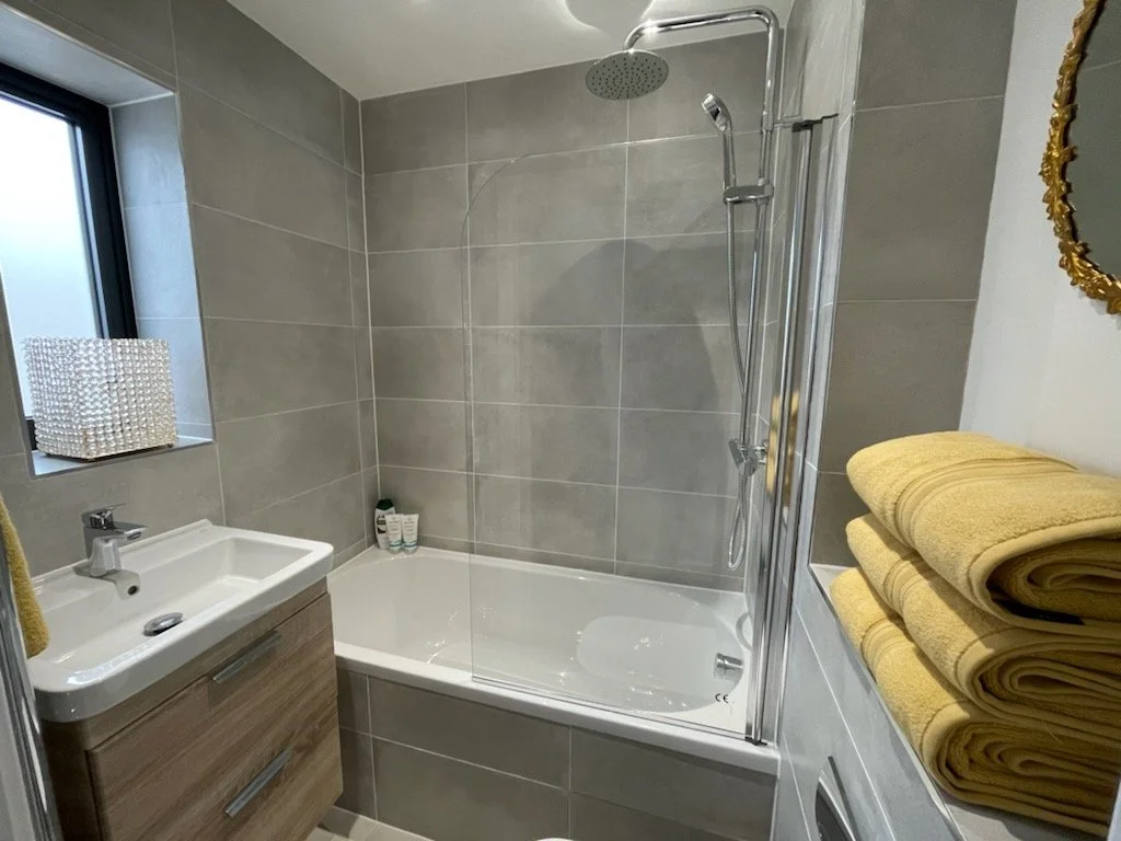 Bathroom with gray tiled walls, a bathtub with glass shower screen, a wall-mounted showerhead, a small sink with a wood cabinet, yellow towels folded on a shelf, and a window with a decorative white cubic container.