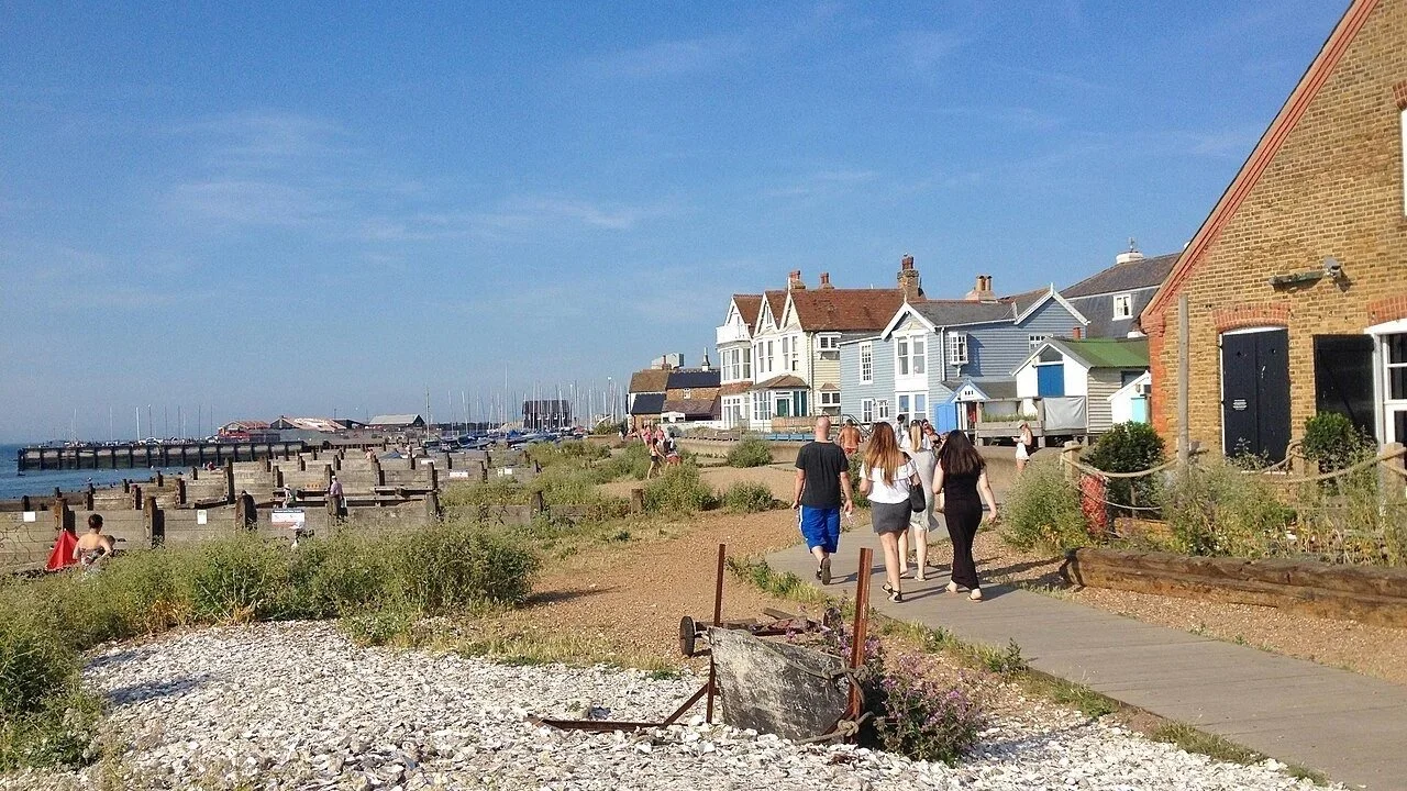 People walking along a coastal boardwalk with houses and sailboats in the background on a sunny day.
