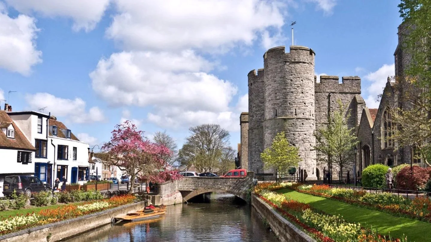 A historic stone castle with rounded turret towers beside a canal with a stone bridge, surrounded by colorful flower gardens and trees, in a small town.