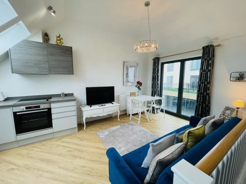 Living room and kitchen area with a blue sofa, a TV on a white stand, a small dining table with chairs, and a sliding glass door leading to a balcony.