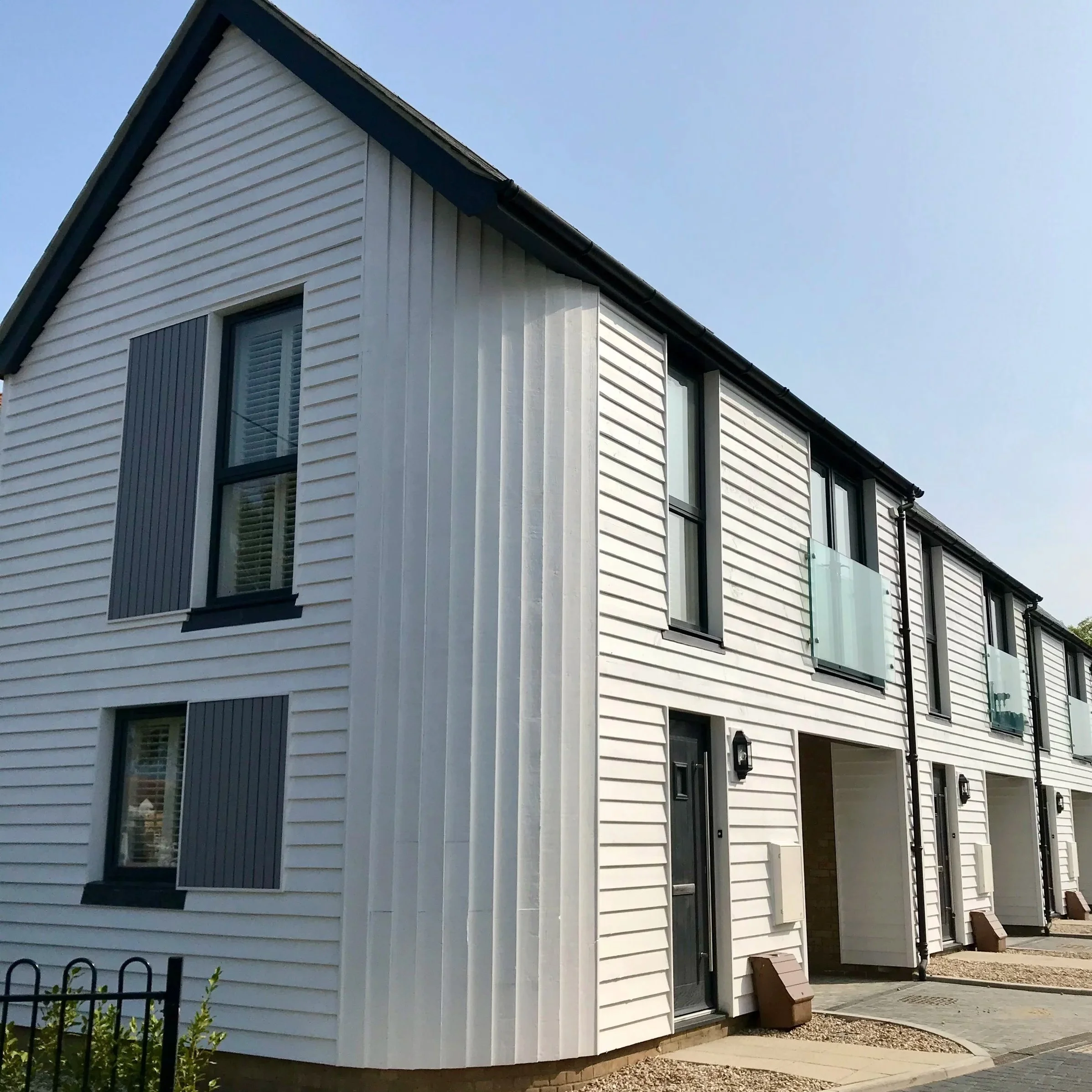 Modern white multi-story building with horizontal siding, large windows, and a sloped roof. The building features dark window trims and a black gutter along the roof's edge. There are some small plants and a bench visible outside the building, with a clear blue sky in the background.
