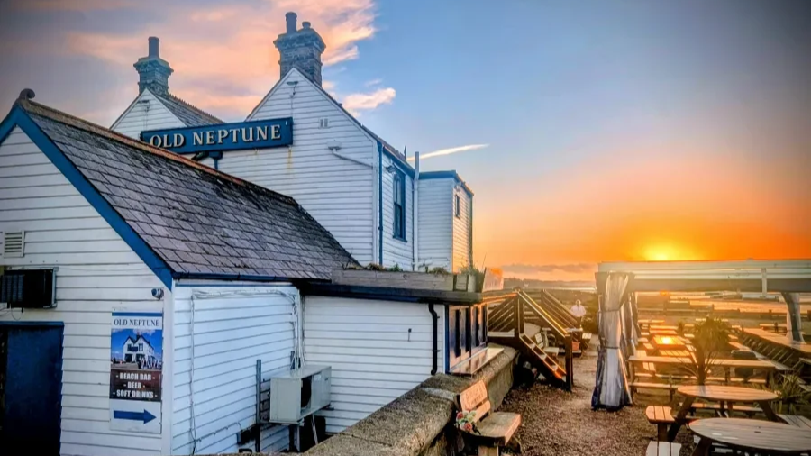 Sunset view over a white seaside building with a sign that reads 'Old Neptune', outdoor seating with wooden tables and chairs, and a staircase leading to an upper level, suggesting it is a beachside bar or restaurant.