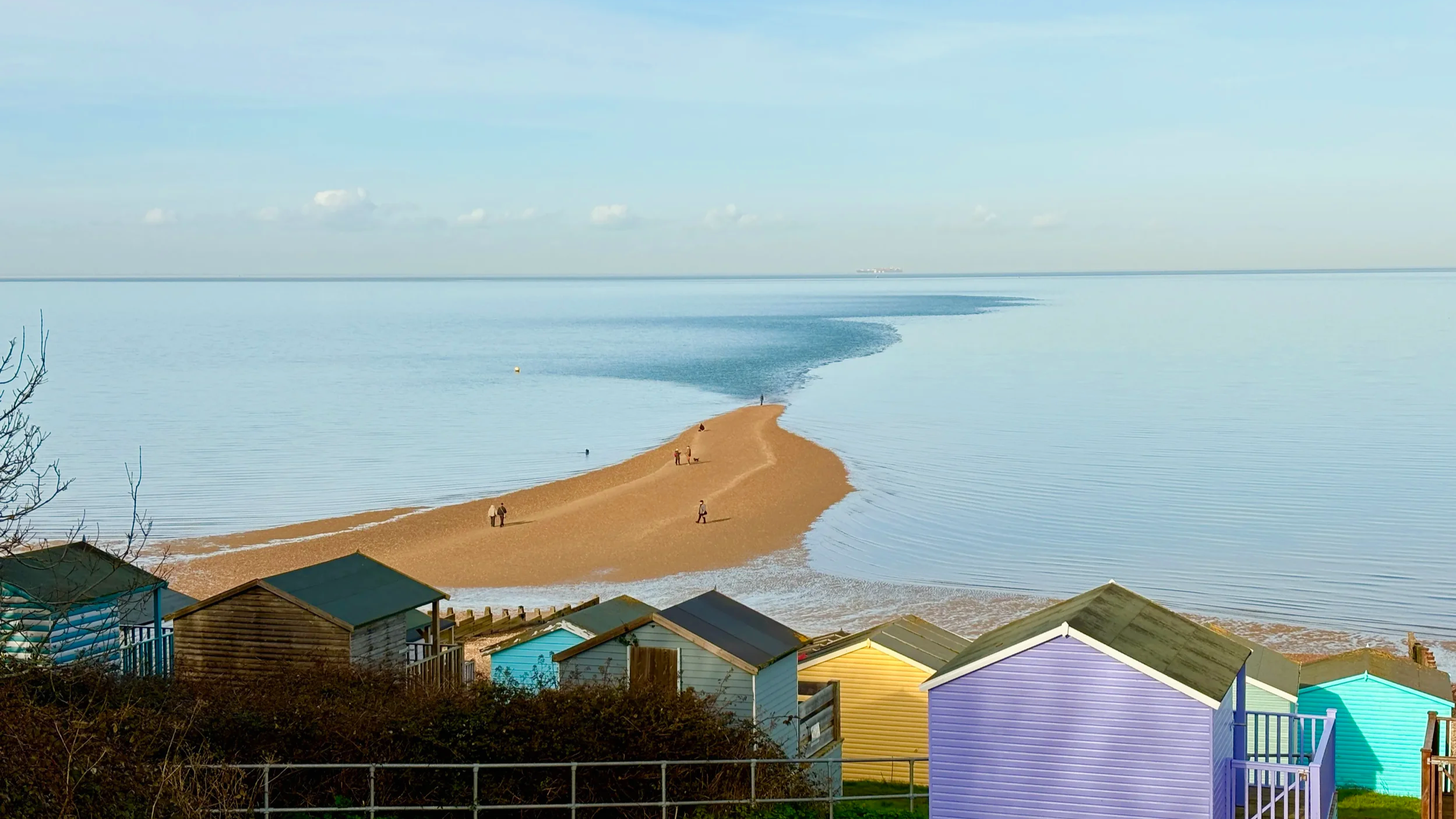 Colorful beach huts and houses on a hillside overlooking a sandy beach and calm water with a small sandbar extending into the water under a cloudy sky.