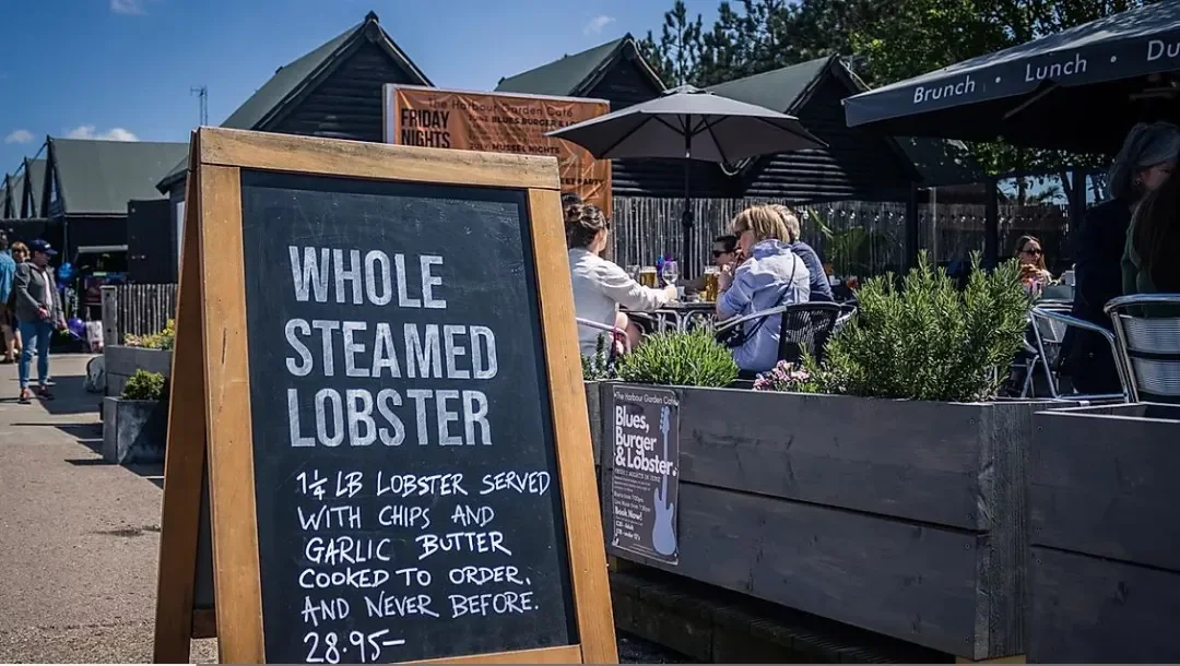 Outdoor restaurant with patrons dining at tables under umbrellas, wooden buildings in the background, and a chalkboard sign advertising whole steamed lobster with chips, garlic butter, for $28.95.