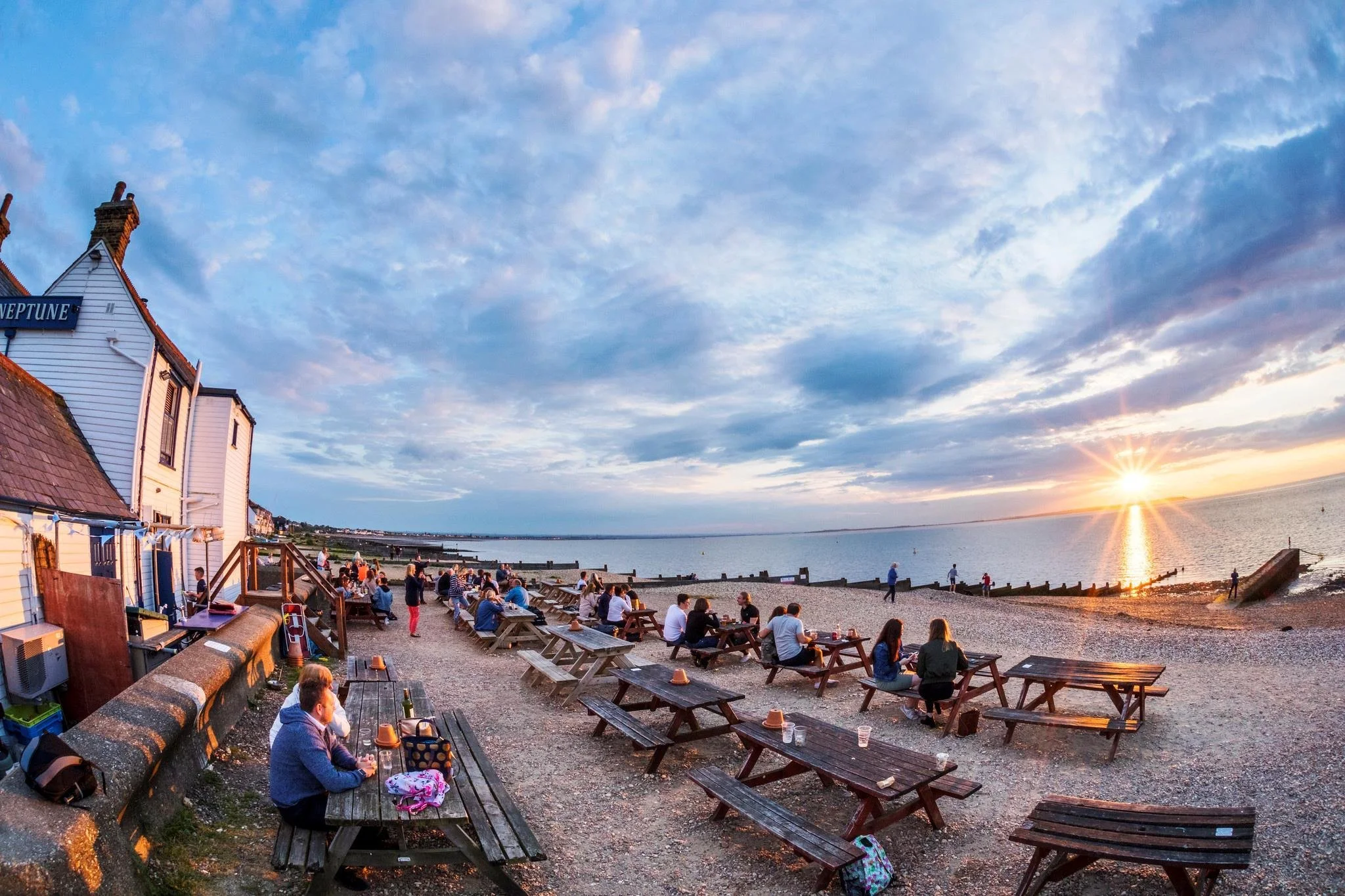 A seaside scene at sunset with a sky filled with clouds and some scattered light, people sitting at wooden benches along a beach promenade, with some walking and others enjoying the view. There are buildings with sloped roofs and a sign on one building that reads 'NEPTUNE'. The water reflects the sunset, creating a warm glow.