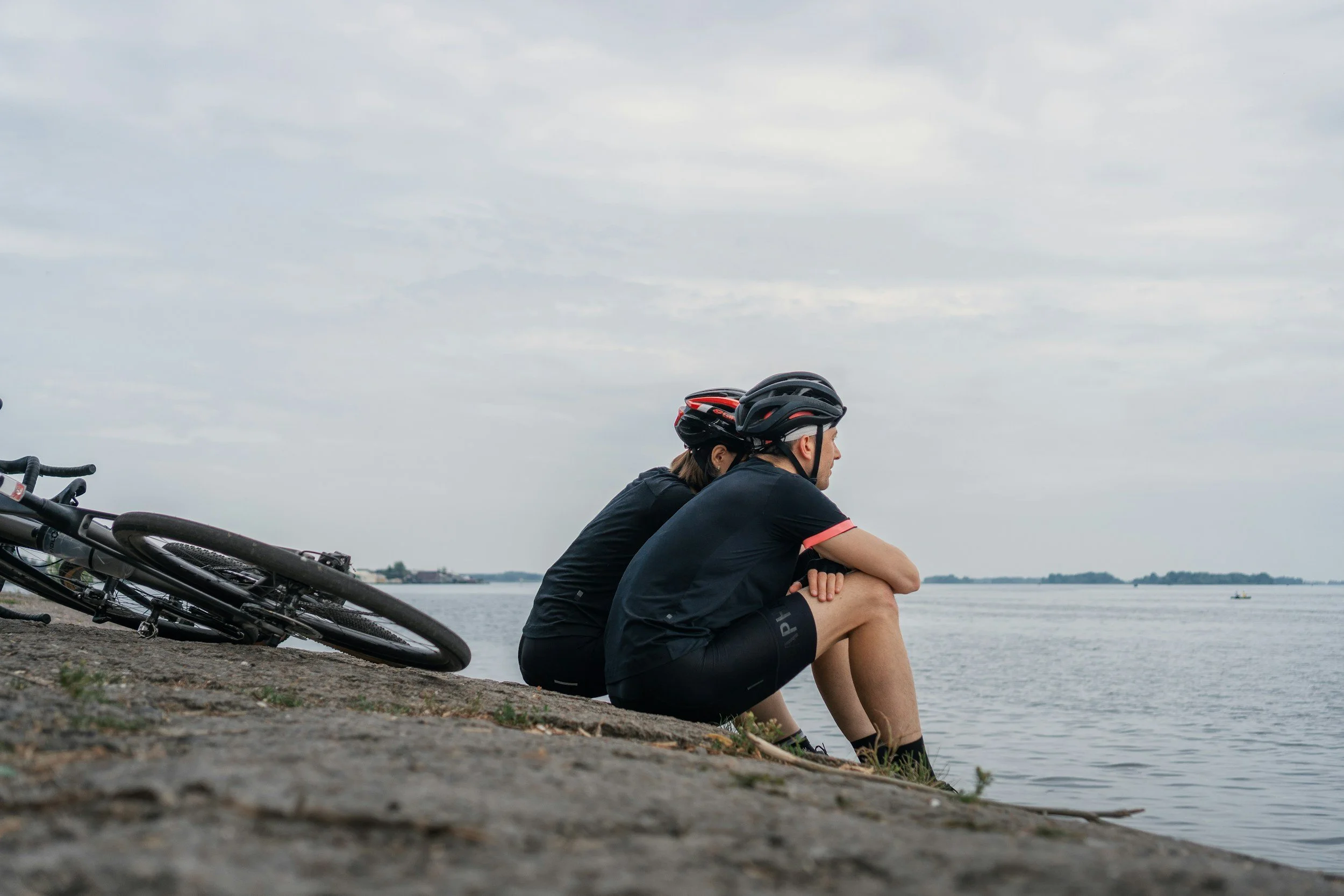 Zwei Radfahrer in Radhelmen sitzen am Ufer eines Flusses, ihre Fahrräder liegen im Sand. Der Himmel ist bewölkt.