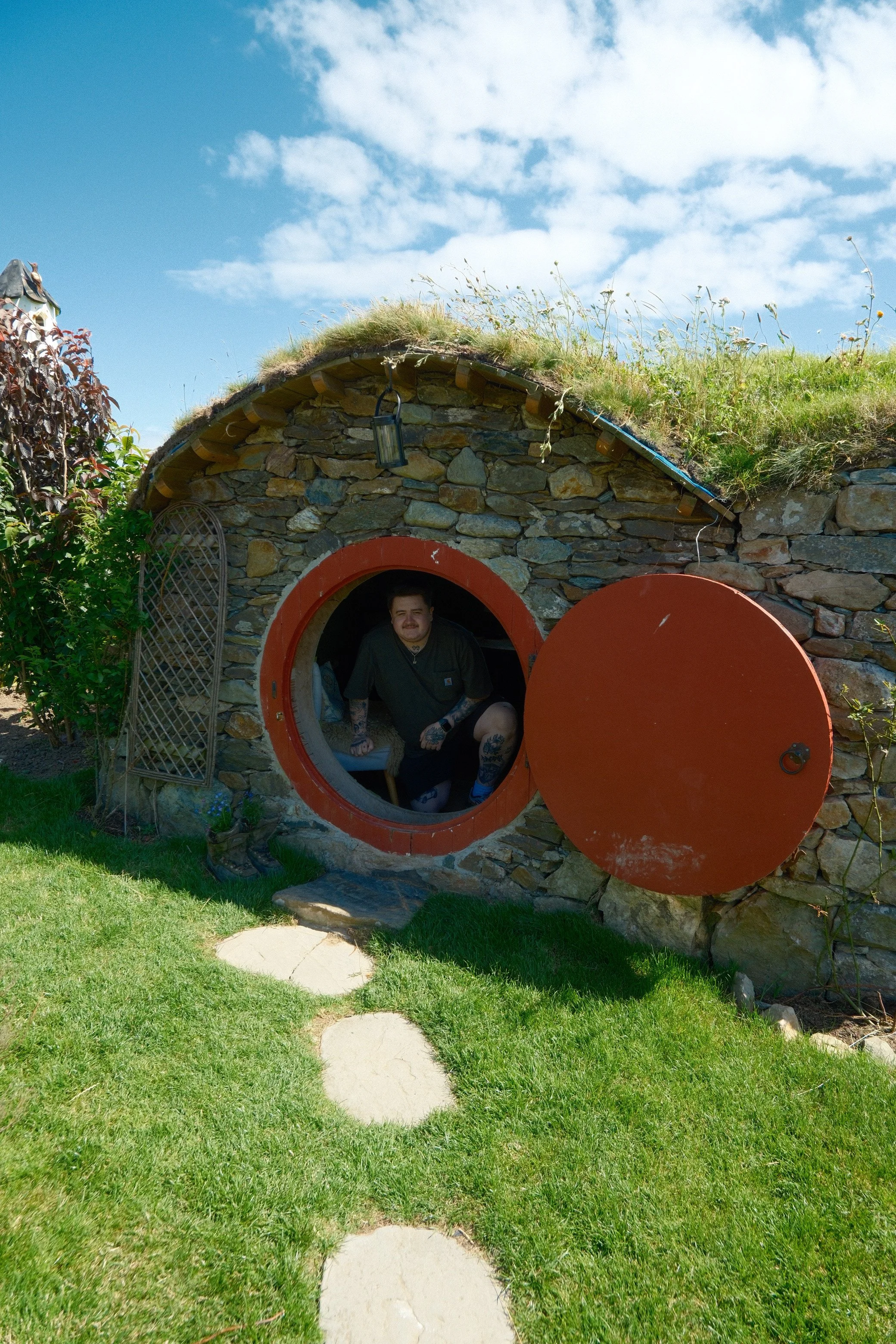 A person going into a small stone house with a round red door in a grassy yard with stone stepping paths, plants, and a blue sky with clouds.