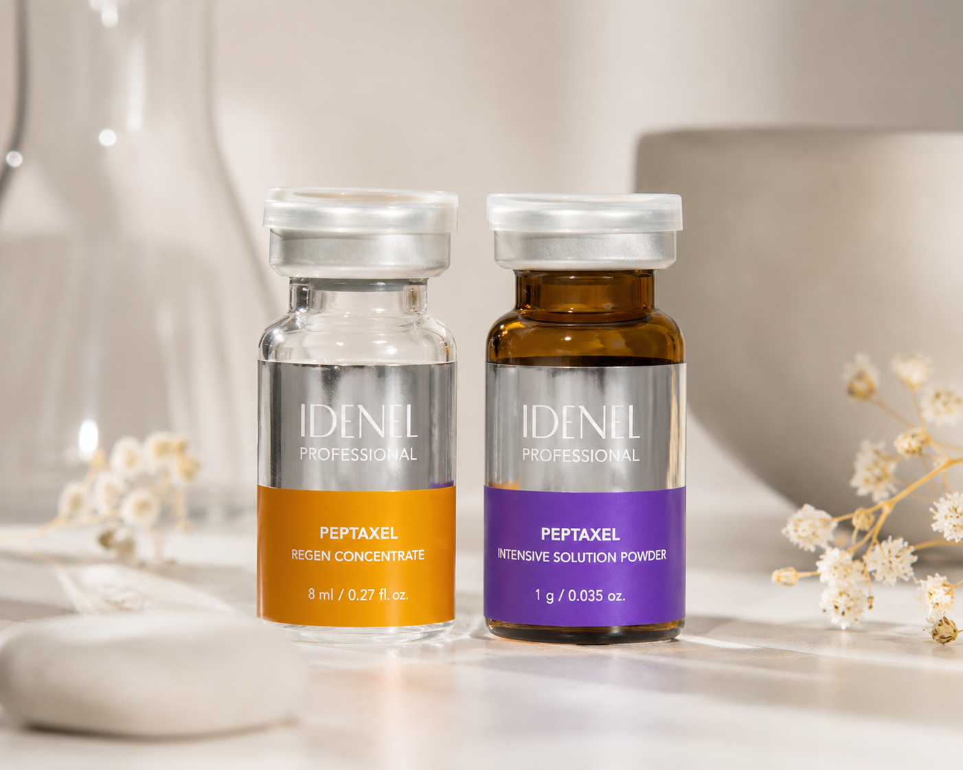Two bottles of IDENEL Professional Peptaxel on a white surface, with a blurred white vase and white flowers in the background.