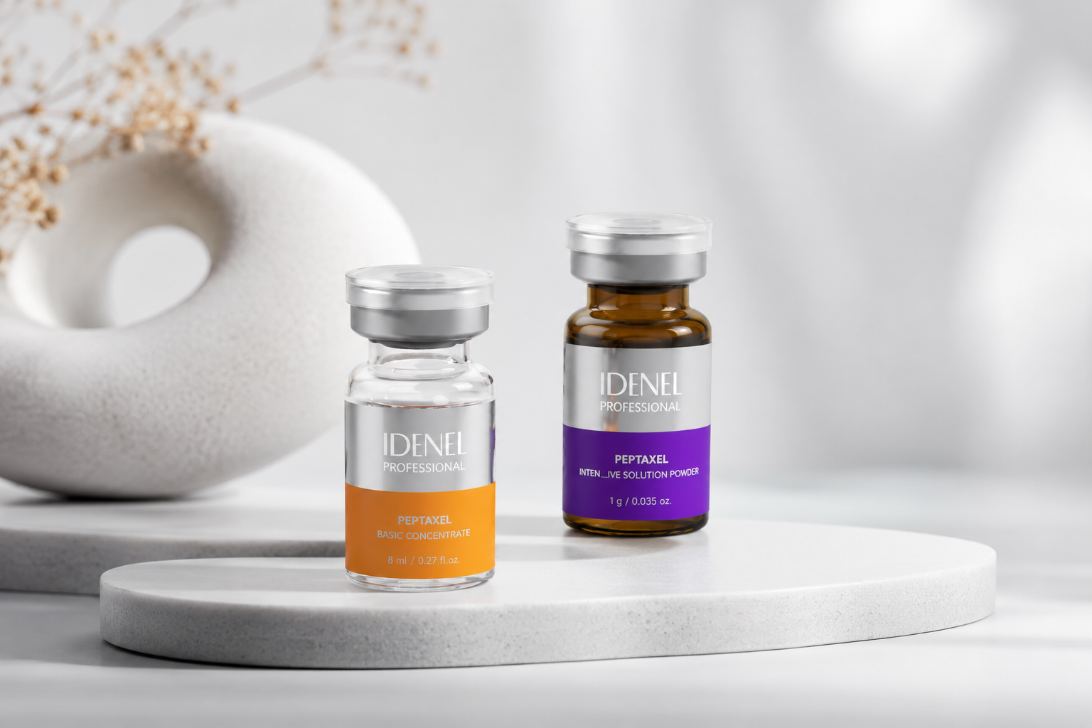 Two bottles of IDENEL Professional Peptaxel skincare products on a white round platform with a textured white vase and dried flowers in the background.