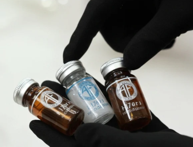 Three small bottles held by hands wearing black gloves, two brown and one clear with a blue label.