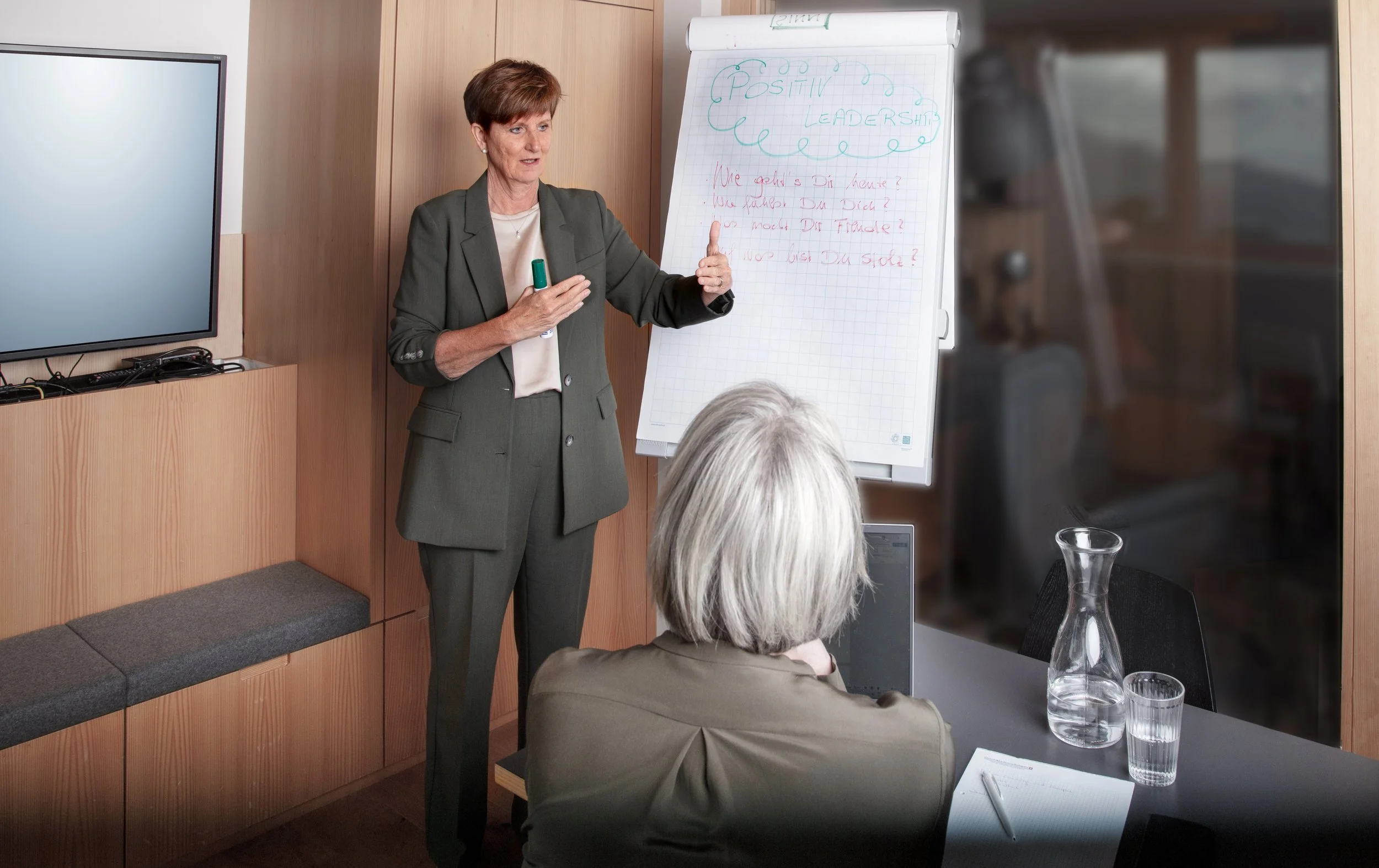 Eine Frau in einem grauen Anzug erklärt vor einer Gruppe in einem Meetingraum. Sie steht neben einem Flipchart mit der Überschrift "Positiv Leadership" und einigen Fragen darunter, während eine ältere Frau mit silbernem Haar, die nur von hinten sichtbar ist, zuhört.