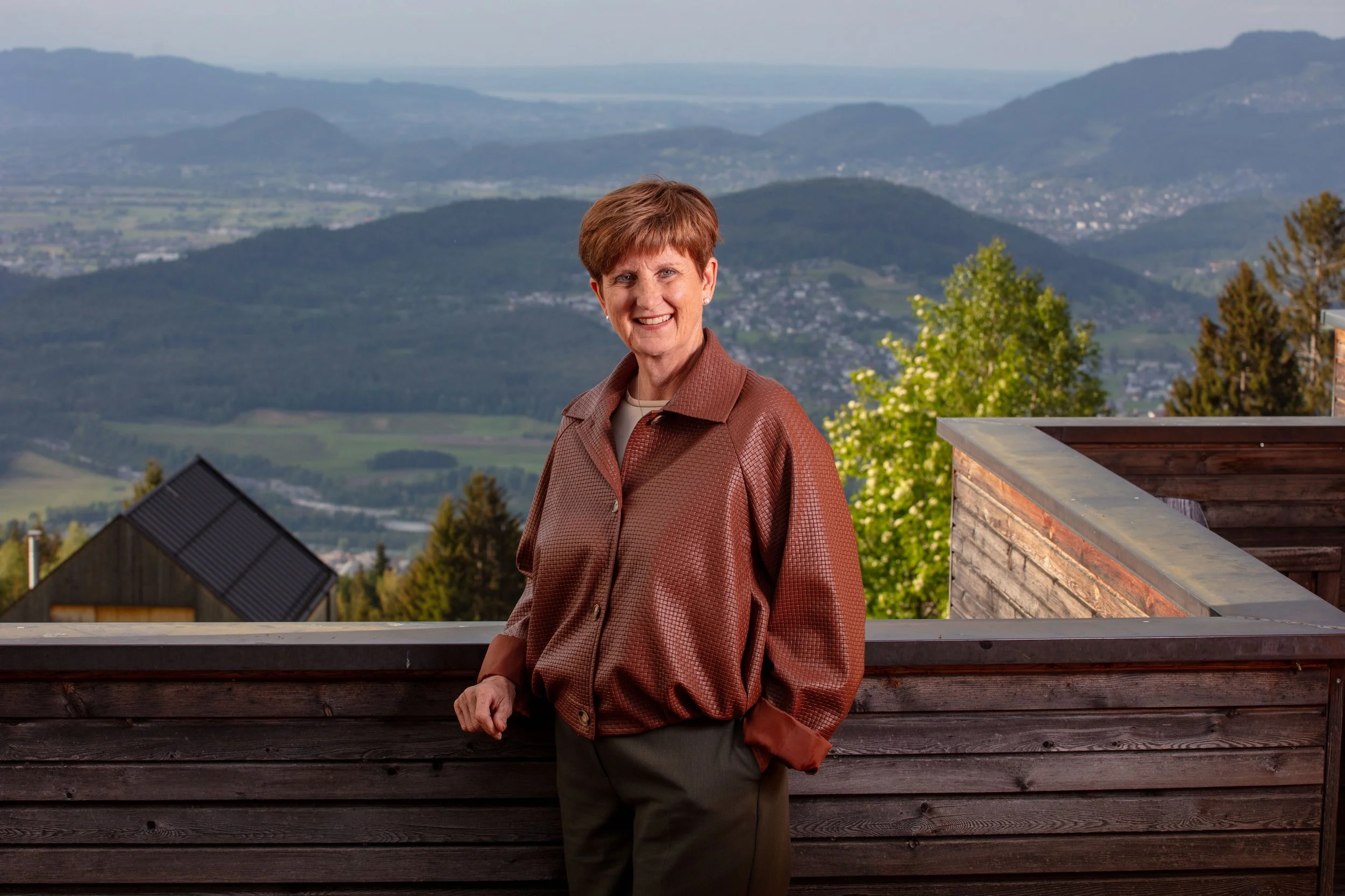 Eine Frau mit kurzen braunen Haaren posiert auf einer Terrasse mit Blick auf grüne Hügel und Berge im Hintergrund. Sie trägt eine braune Jacke und ein helles Oberteil. Die Szenerie wirkt ruhig und malerisch.