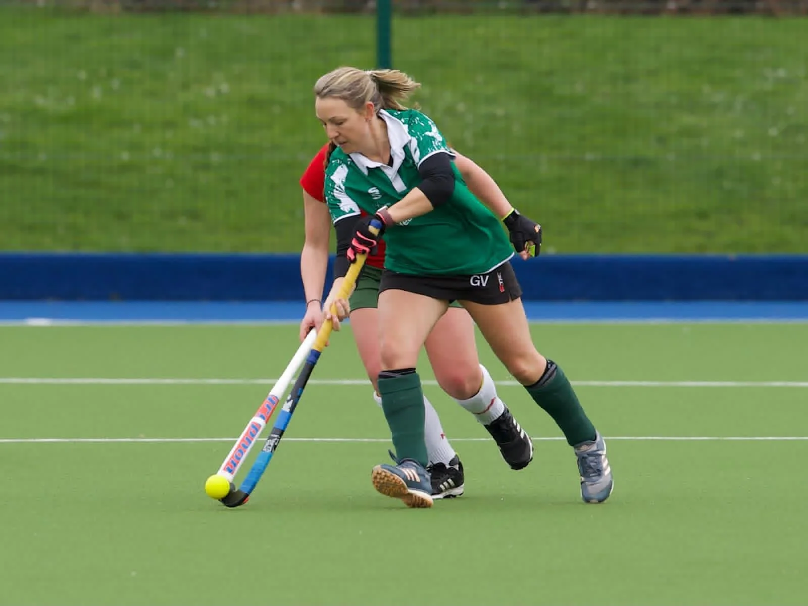 Two women playing field hockey on a green turf, with one woman in a green and black uniform and the other in a red and green uniform, competing for the ball.