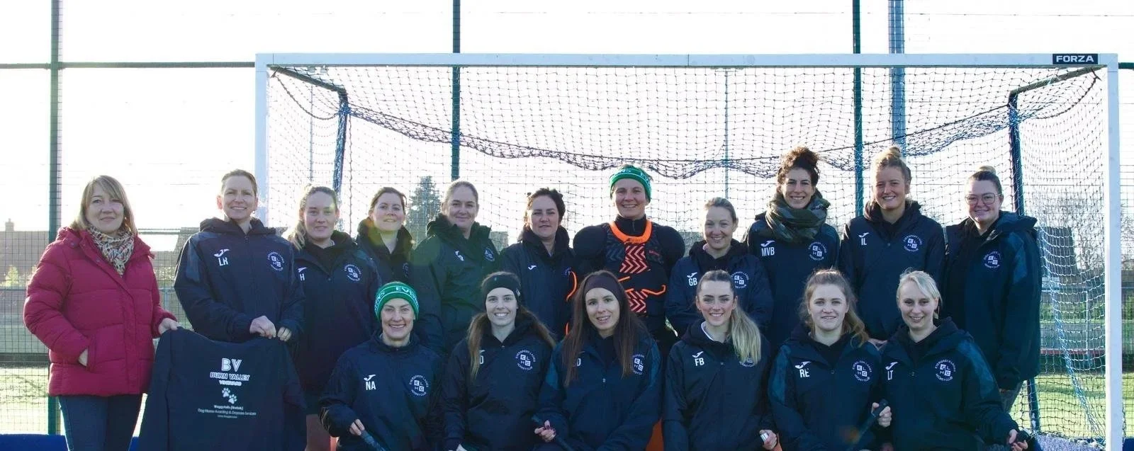 Group of female athletes and coaches standing in front of a soccer goal on an outdoor field, smiling for a team photo.