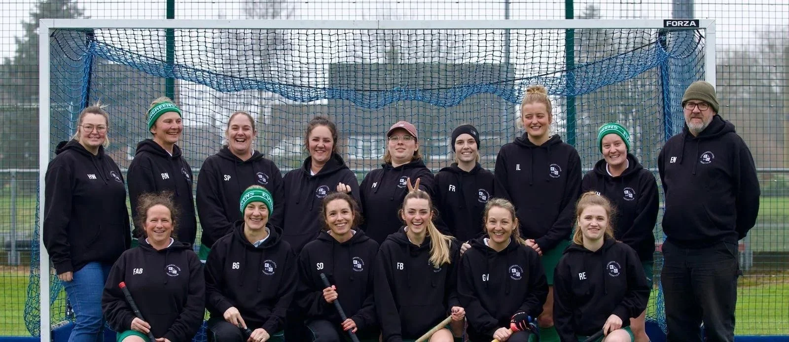 A group of women and one man standing in front of a field hockey goal on a sports field, all dressed in black hoodies with team logos, smiling for a team photo.