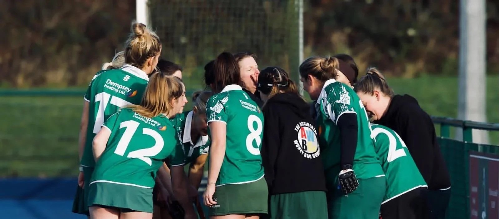 Group of female field hockey players in green uniforms gathered around their coach, listening during a game.