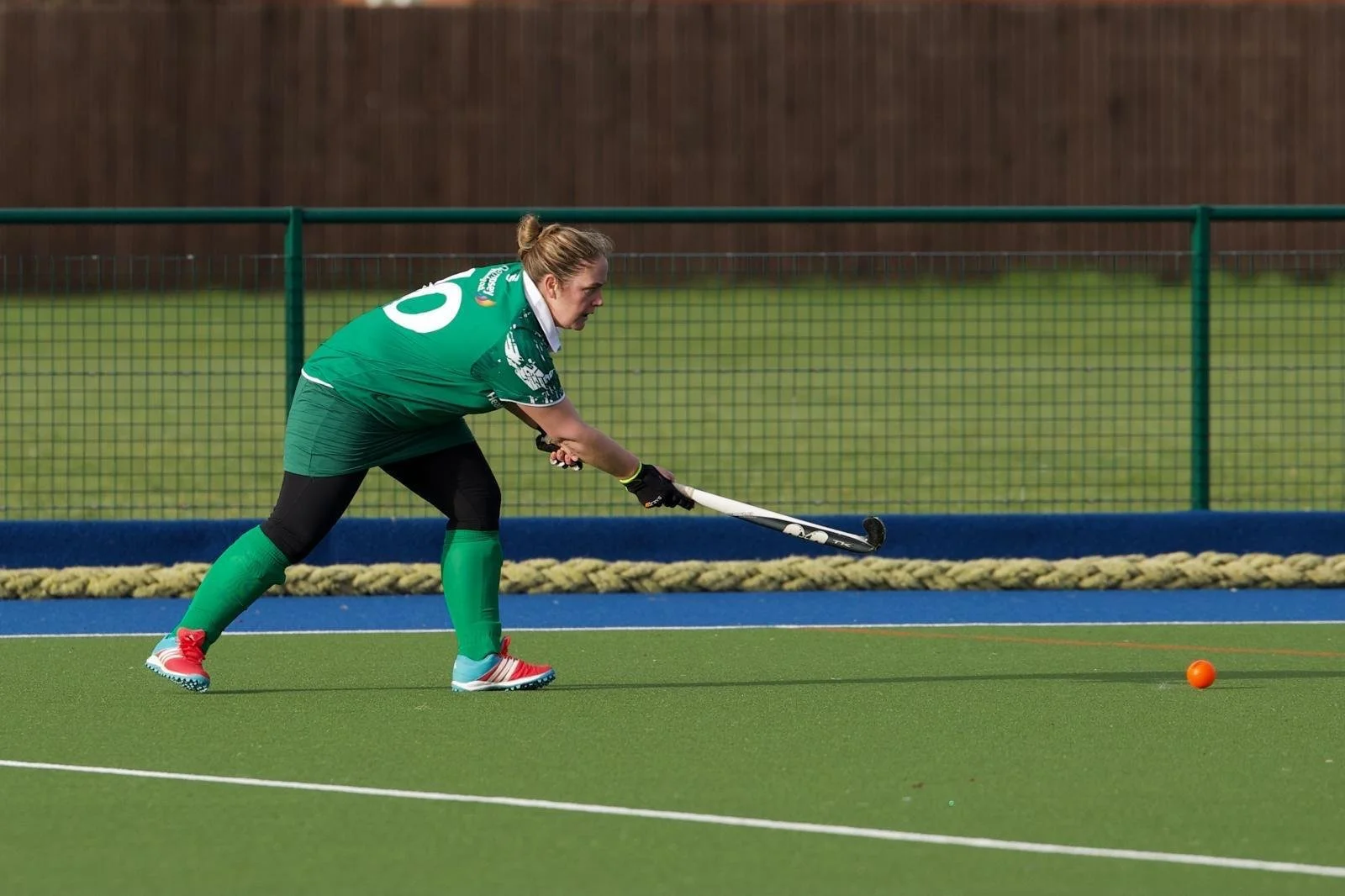 A female field hockey player in a green uniform, bent forward, holding a hockey stick, with an orange ball on a green field, near a blue hockey turf line, and a green fence in the background.