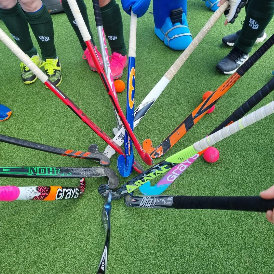 A group of field hockey sticks arranged in a circle on a green artificial turf surface, with some children standing around in athletic shoes.