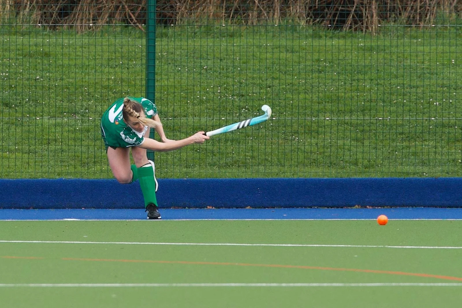 A female field hockey player in green uniform is attempting to hit an orange ball with her stick on a green and blue field, with a fence and trees in the background.