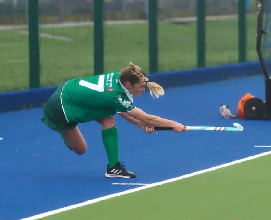 A female field hockey player in a green jersey and shorts, bending forward with her stick, on a blue field with green fencing in the background.