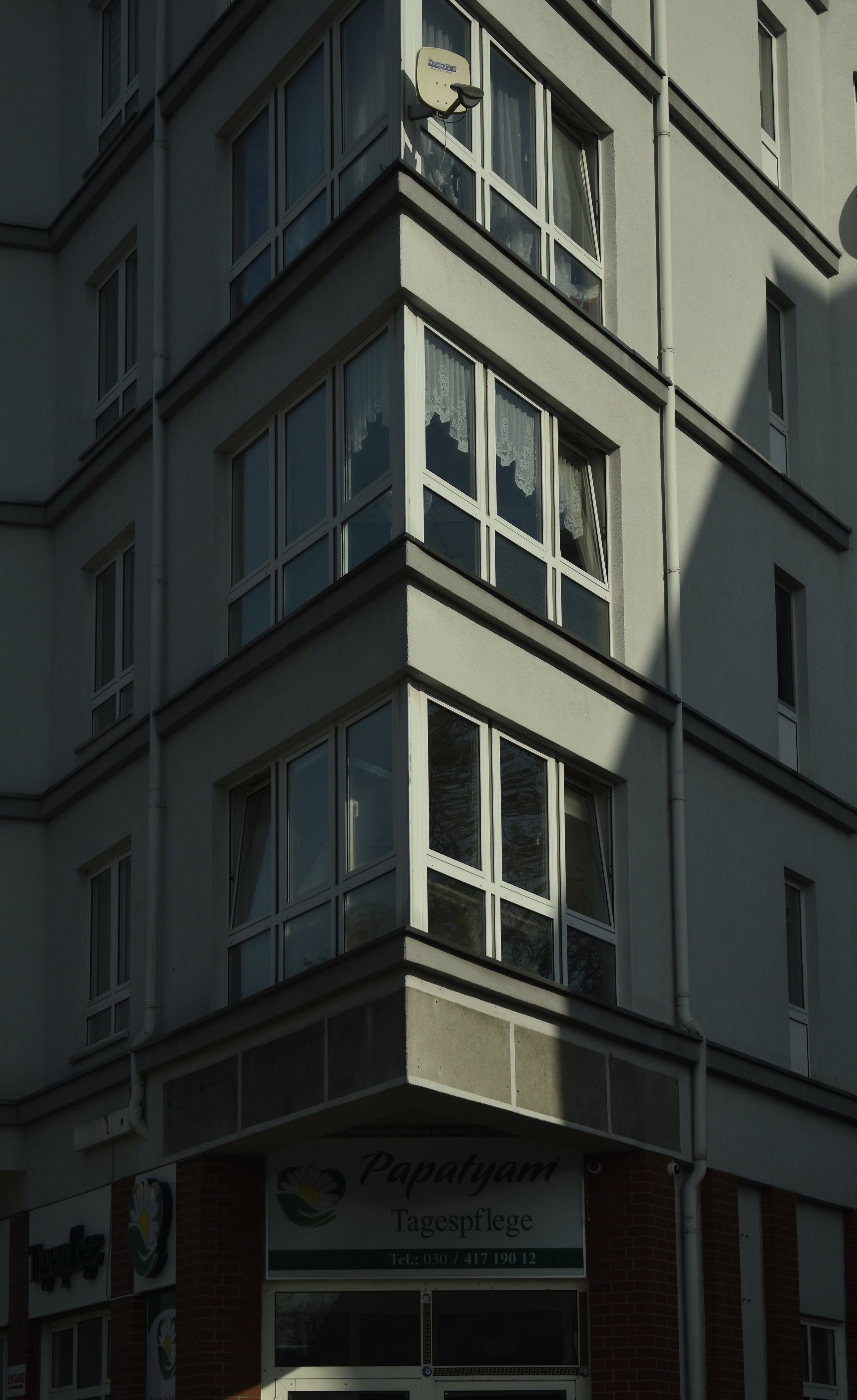 A corner of a multi-story building with large windows, some with lace curtains, and a satellite dish mounted on the upper part of the building. The lower part of the building has signs for a business called Papatyam Tagespflege with a phone number.