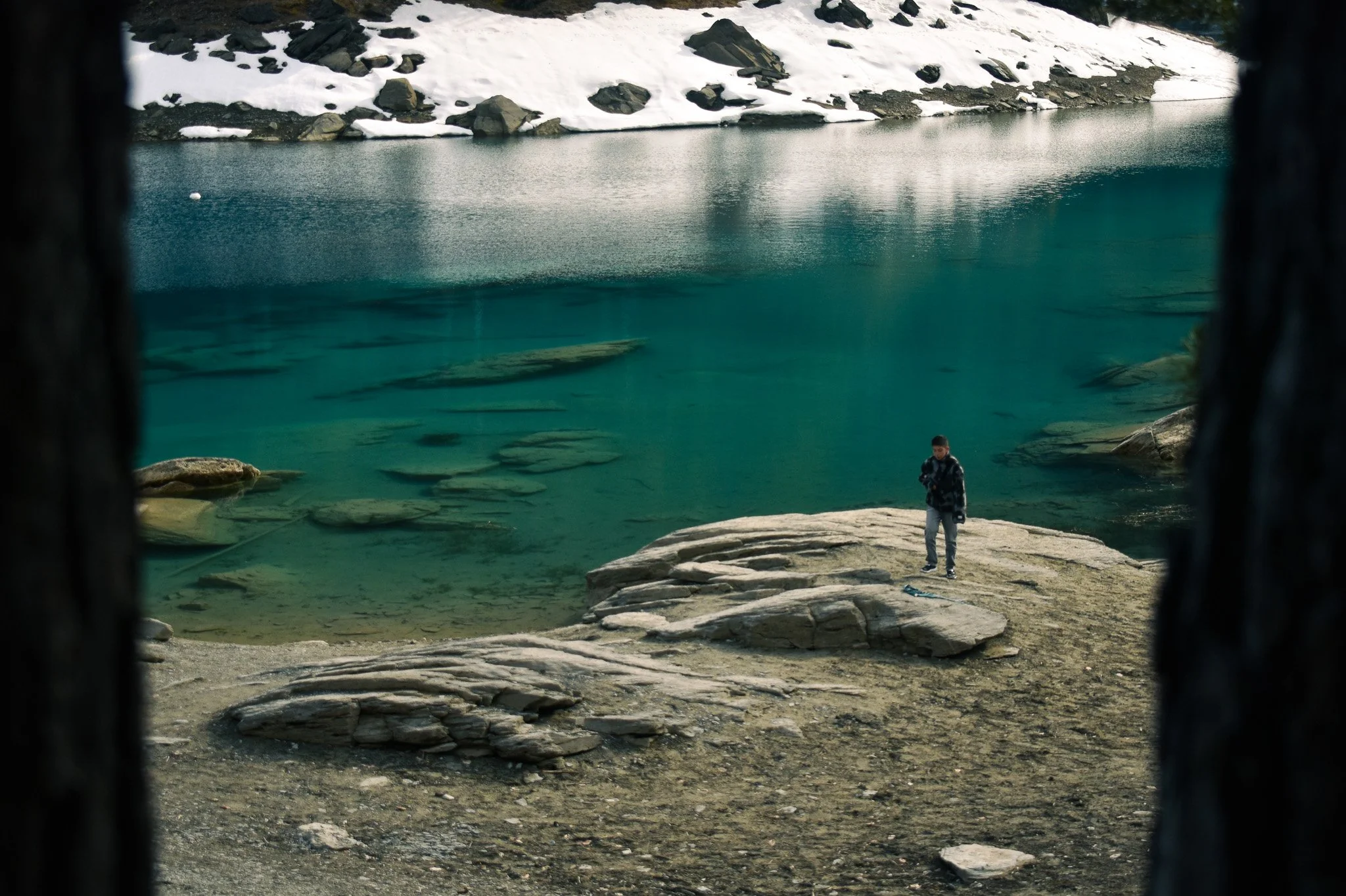 A person standing on a rocky shoreline near clear turquoise water, with snow-covered rocks and hills in the background, viewed through a framed opening.