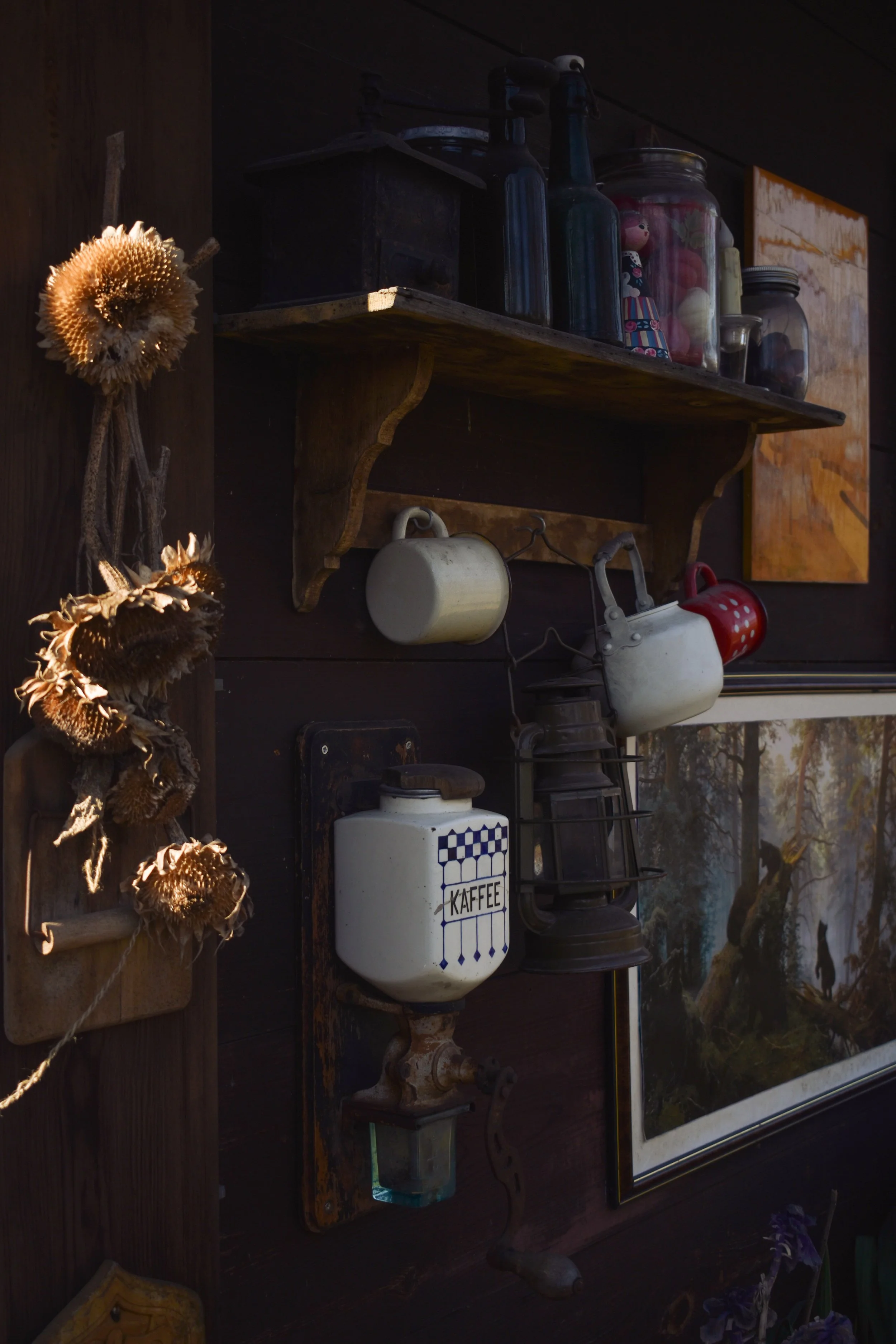 Wall decorated with dried flowers on the left, a wooden shelf with glass jars, bottles, a small doll, and old kitchenware, including a vintage coffee container with a checkered pattern and the word "KAFFEE" on it. A framed painting of trees in a natu