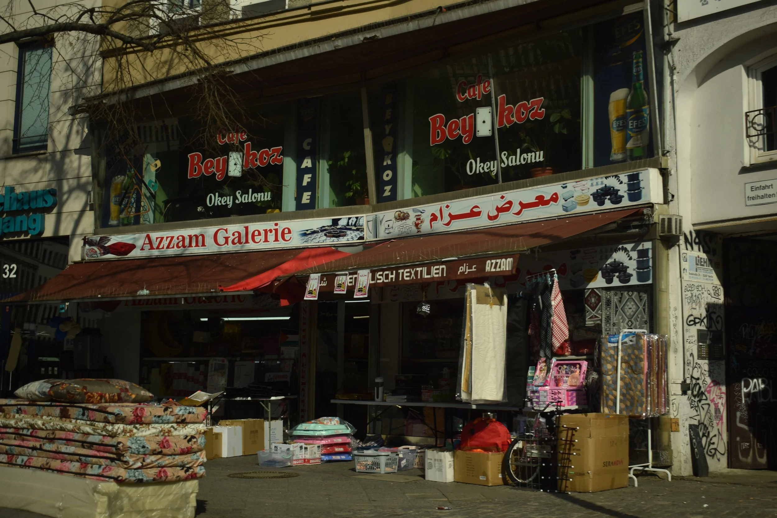 Street scene with a store selling various household items and textiles. Sign on the upper storefront reads 'Café Beykoz' and 'Azzam Galerie' with additional signs in Arabic and Turkish. Items like mattresses, boxes, and shopping baskets are displayed
