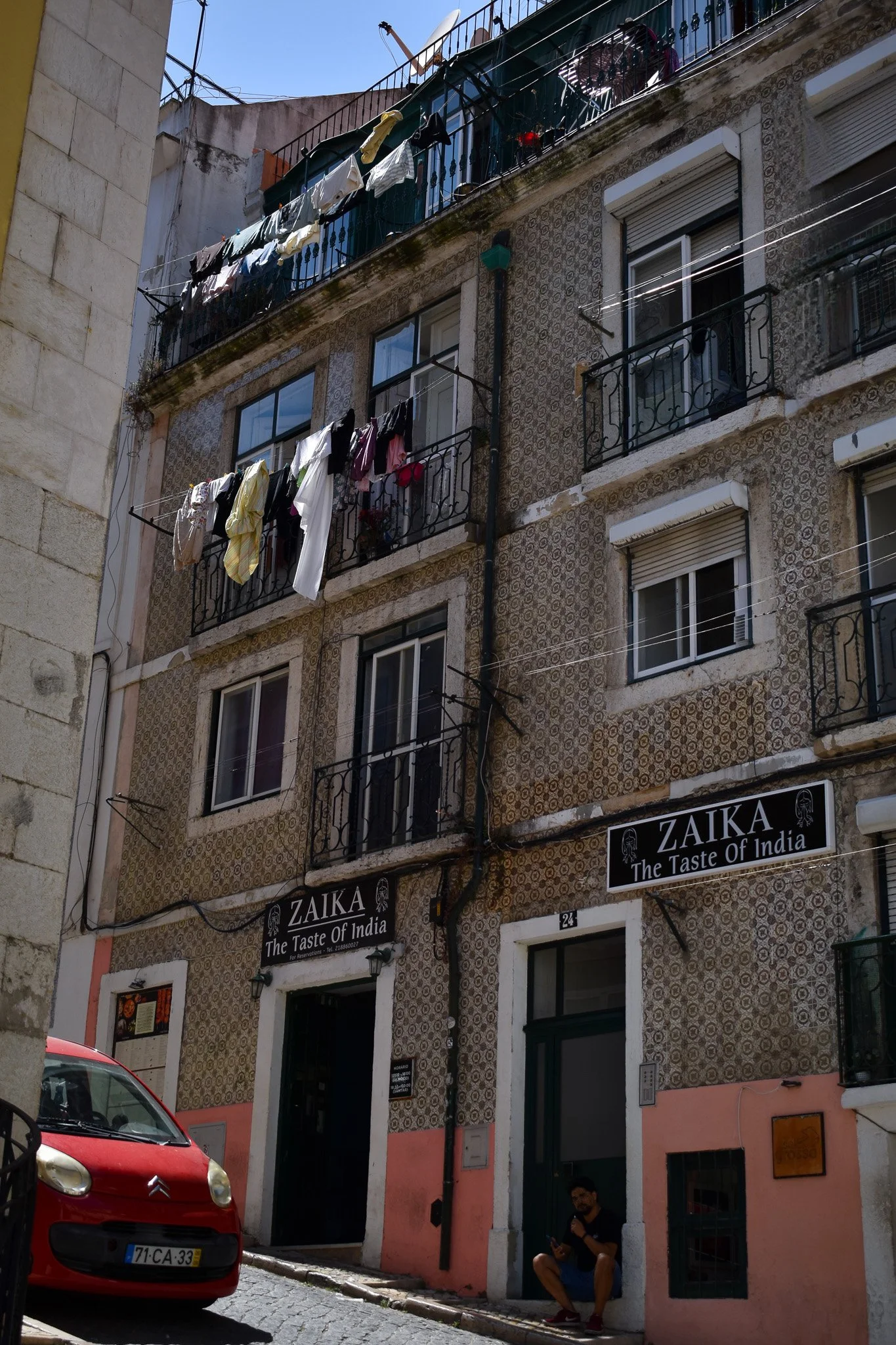 Street view showing a building with outdoor laundry hanging on balconies, a red car parked on the cobblestone street, and a man sitting on the sidewalk in front of a restaurant named ZAIKA, The Taste of India.